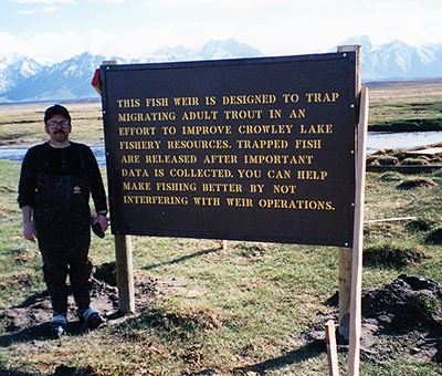 Sam Bertoni standing next to the Owens River weir construction informational signage 1990 This fish weir was constructed by the Izaak Walton League under the direction of Sam Bertoni