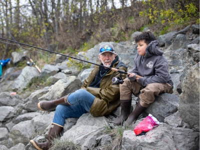 An older man sitting on a rocky stream edge with a little boy showing him how to fish.