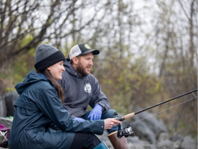 A man sitting on the edge of a stream with a woman showing her how to fish