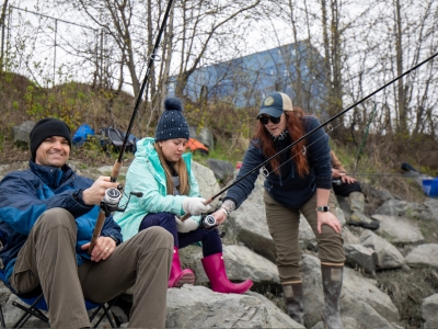 A man fishing next to a woman holding a fishing rod and another woman showing her how to fish on the edge of a river